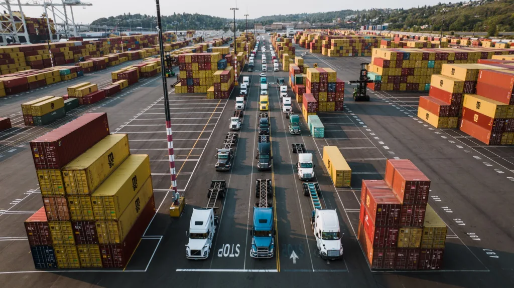 Semi trucks driving through a large container shipping yard with stacked cargo containers at a busy freight port