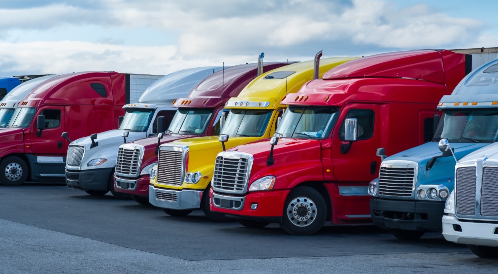 A lineup of colorful semi-trucks, primarily red, with a yellow and a white truck parked in a lot under a partly cloudy sky.